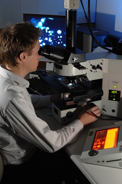 A man sitting at a microscope with a screen in the background showing oil inclusions