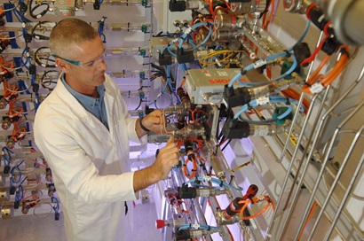 A male scientist in lab coat and safety glasses standing among racks of components