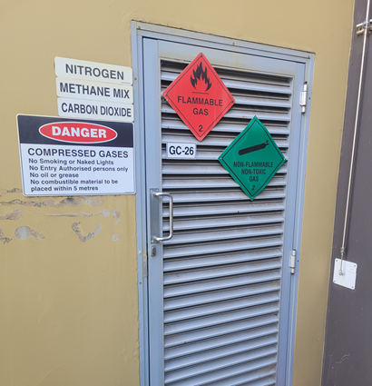 A metal louvered door on a building, displaying multiple safety signs indicating the presence of hazardous gases. On the door are two diamond‑shaped hazard signs: a red “Flammable Gas – 2” sign with a flame symbol, and a green “Non‑Flammable, Non‑Toxic Gas – 2” sign with a gas cylinder symbol. To the left of the door are additional labels listing gases stored inside: Nitrogen, Methane Mix, and Carbon Dioxide. Below these is a large warning sign reading “Danger: Compressed Gases” with instructions including no smoking, no naked lights, authorised entry only, no oil or grease, and no combustible materials within five metres.