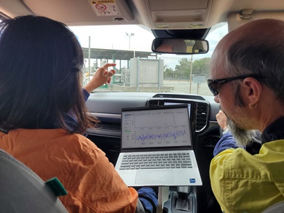 Two people are seated inside a vehicle reviewing data on a laptop. The laptop screen shows graphs and measurement readings. One person gestures toward equipment visible outside the vehicle, seen through a fenced industrial area.
