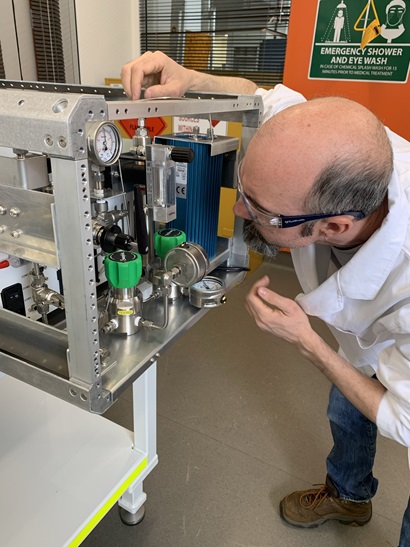 Person in a laboratory setting inspecting a metal equipment unit with gauges, valves and tubing. They are leaning in closely while adjusting a component on the top of the device. Safety signage, including an emergency shower and eye‑wash sign, is visible in the background.