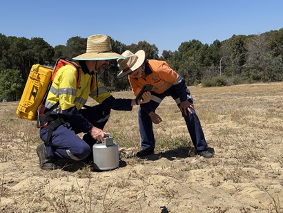 Two workers in high‑visibility clothing are taking measurements in a dry field. One is kneeling beside a small metal sampling chamber on the ground, while the other crouches nearby checking a handheld device. Trees and clear sky are in the background.