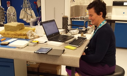 teacher sitting at tabel with laptop and ipad in front and acoustic testing materials at the back of bench