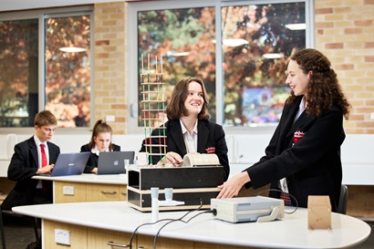 Two high school students working with equipment in a classroom with two other students working on laptops in the background