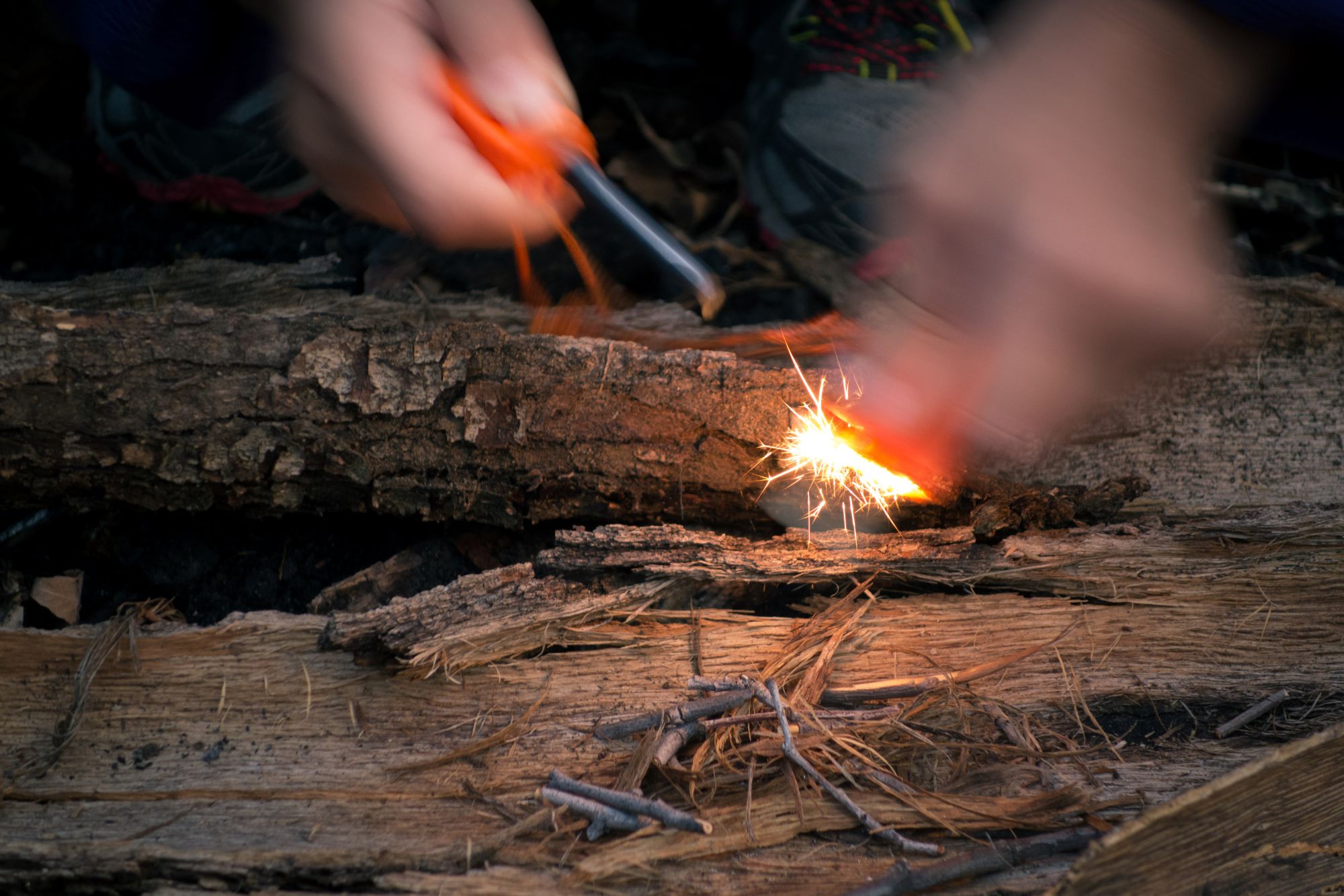 Two blurred hands generating sparks over brown dry logs to start a fire.