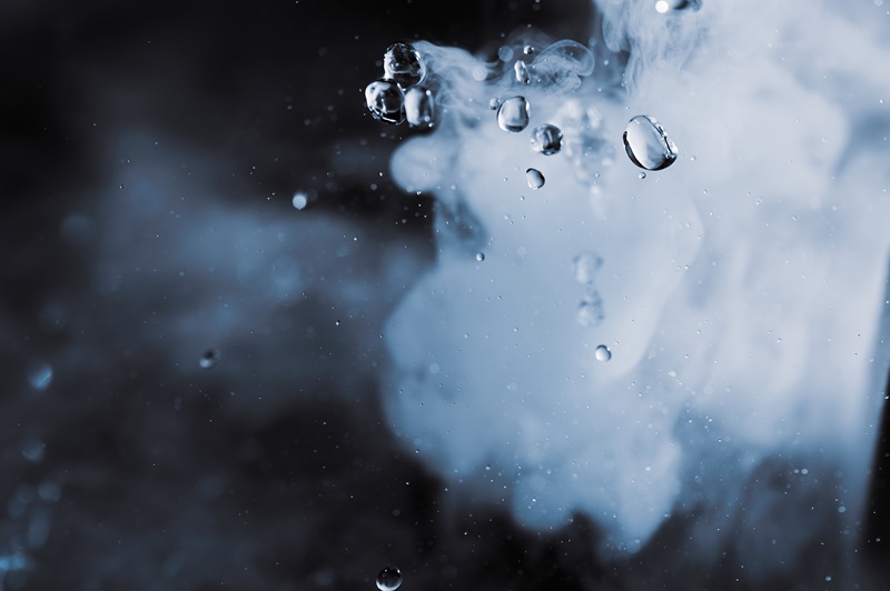 Photograph showing water droplets suspended in air with a background of swirling white smoke or mist against a dark backdrop. 