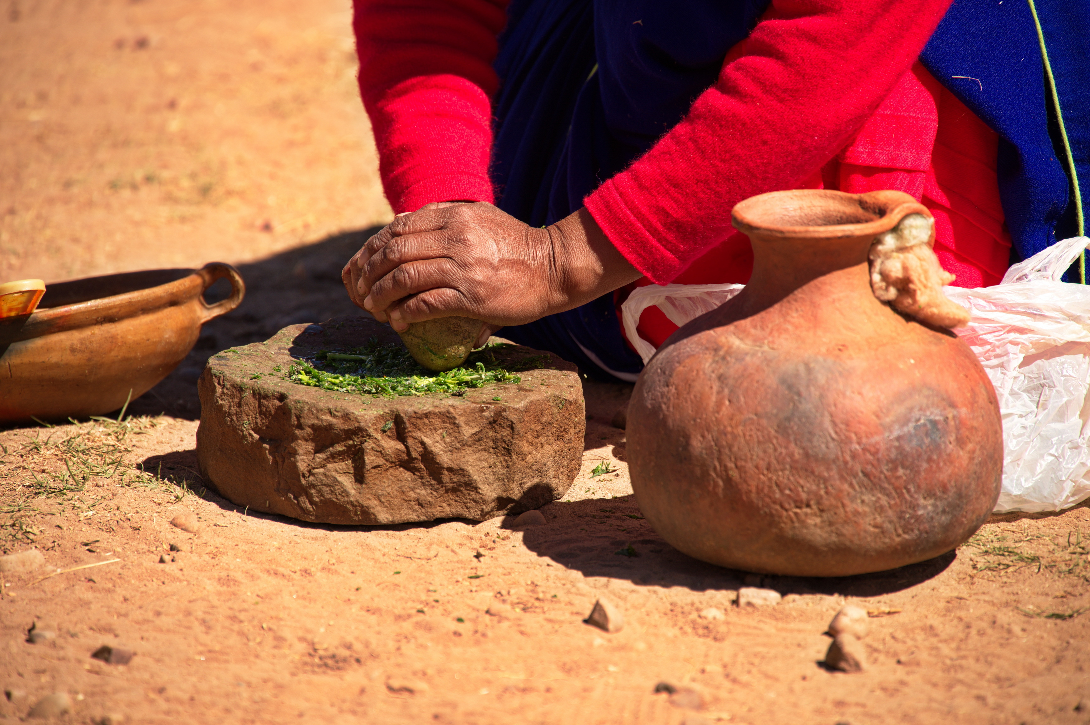 Photograph showing a person using a traditional stone grinder to crush green herbs on a dirt ground. Surrounding the grinder are rustic clay pots, highlighting a cultural or historical food preparation method.