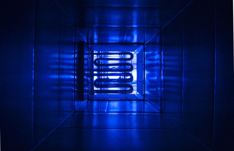 Photograph showing the interior of a metallic duct illuminated by blue light, highlighting a coiled heating element at the far end. The image depicts the internal heating element from a CSIRO-designed climate testing rig.