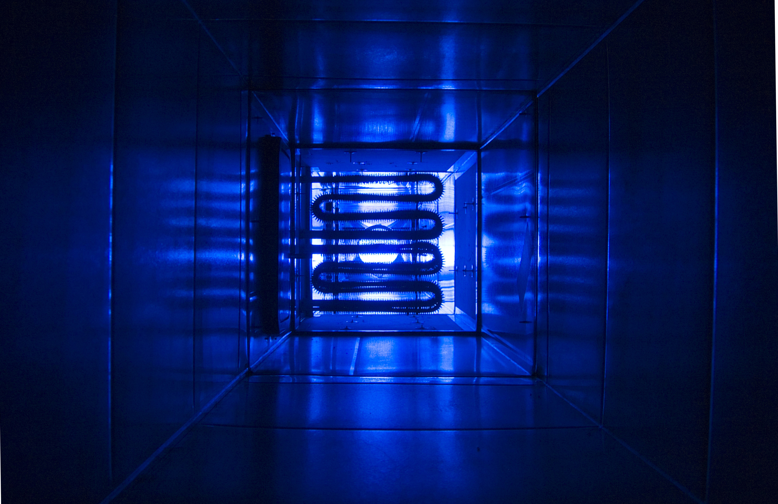 Photograph showing the interior of a metallic duct illuminated by blue light, highlighting a coiled heating element at the far end. The image depicts the internal heating element from a CSIRO-designed climate testing rig.