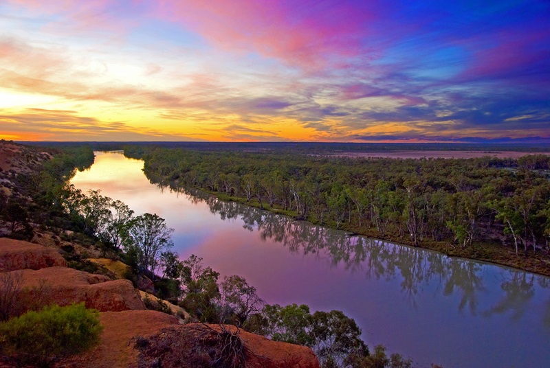 Photograph of a river winding through a dense forest under a colorful sunset sky with shades of pink, purple, and yellow. The image is of the Murtho floodplain in the Murray-Darling Basin, near Renmark, South Australia.