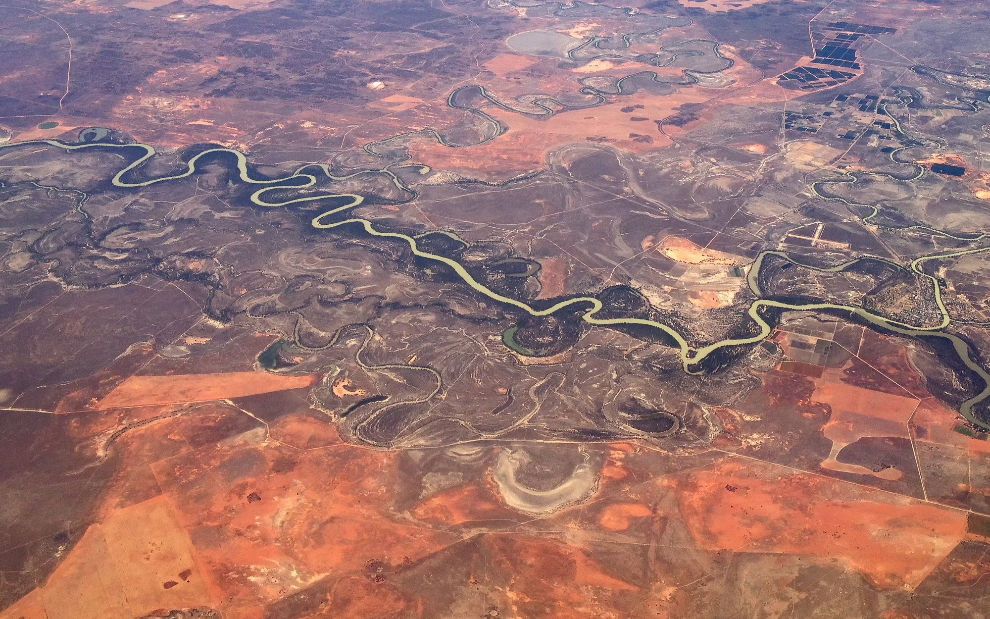 Aerial photograph showing a winding river cutting through a dry, arid landscape with reddish-brown soil and sparse vegetation. The scene depicts the River Murray downstream of the junction with the Darling River at the town of Wentworth (far right).