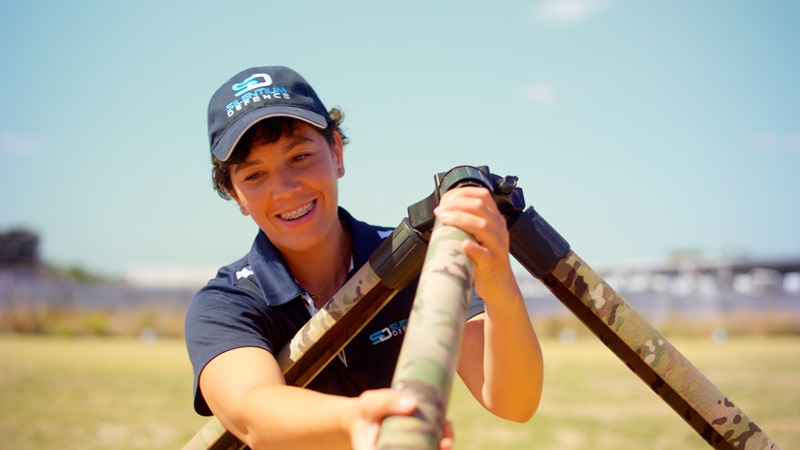 Photograph of a person outdoors holding two camouflaged cylindrical objects crossed in front of their chest, wearing a navy blue shirt and matching cap with a logo. Background shows an open field with blurred structures and clear sky.
