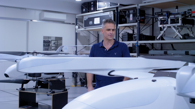 Photograph of a person standing behind multiple white drone-like unmanned aerial vehicles (UAVs) in a storage or workshop setting.