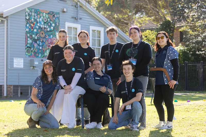 A group of students and mentors, some kneeling on the ground and others standing, smiling at the camera.