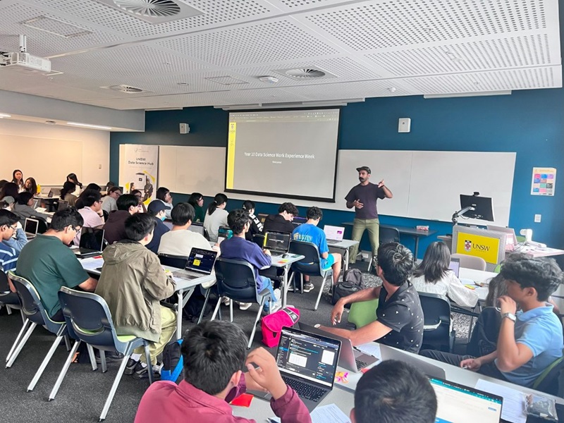Students sit at desks using laptops during a UNSW Data Science Work Experience Week session, while a facilitator leads a lesson at the front with a projected presentation.