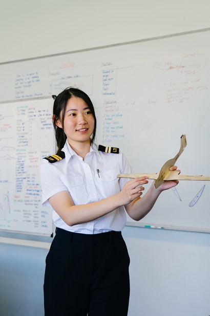 Person in a pilot uniform holding a model glider while teaching in front of a whiteboard filled with diagrams and notes.