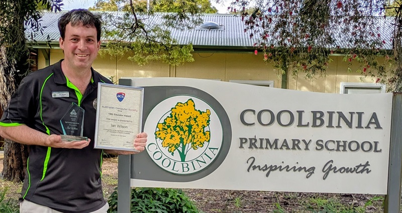 An man stands beside the Coolbinia Primary School sign holding a trophy and framed award certificate recognising achievement in digital technologies education.