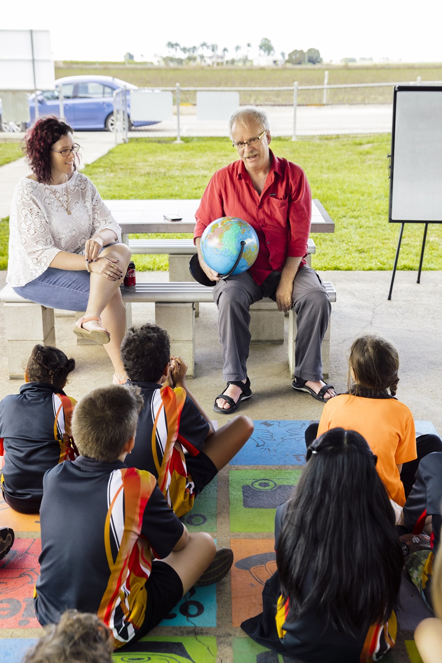 STEM professionals sitting outside at a bench teaching a group of children sitting on the ground. 