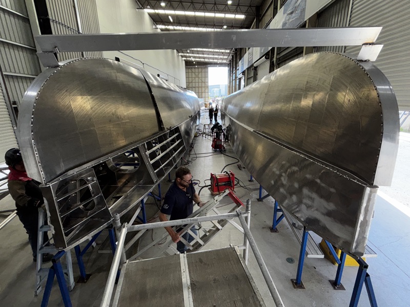Two hulls under construction in warehouse, with workers around.