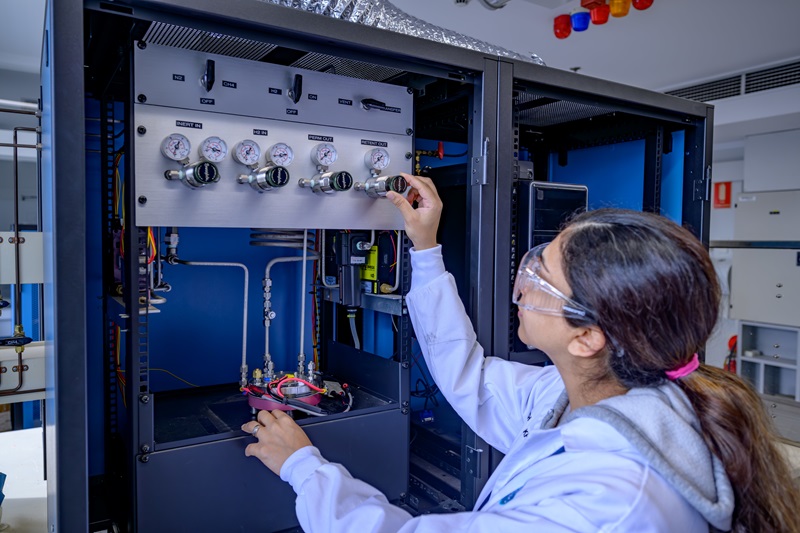 A woman in a lab coat and glasses operates a piece of machinery by turning a dial. 