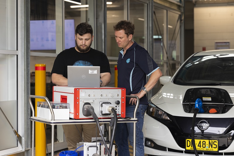 Two men stand in a warehouse working on a laptop, configuring a machine connected to an electric vehicle.