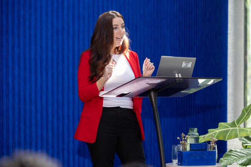 Picture of woman in red jacket at lectern speaking to an audience (the audience cannot be seen).