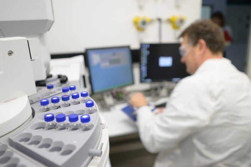 Research equipment in the foreground, man in white coat in background working on computer.
