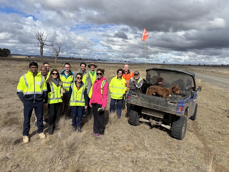 Group of researchers stand smiling at the camera in a field on a farm, with high vis jacket on, with a buggy and a dog on the back next to them. 