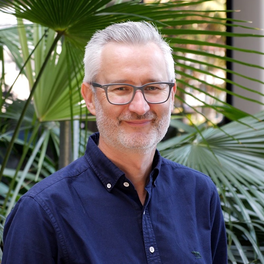A man with short grey hair and thin-framed glasses stands against a backdrop of fern fronds. He is wearing a navy blue shirt and smiling slightly. 