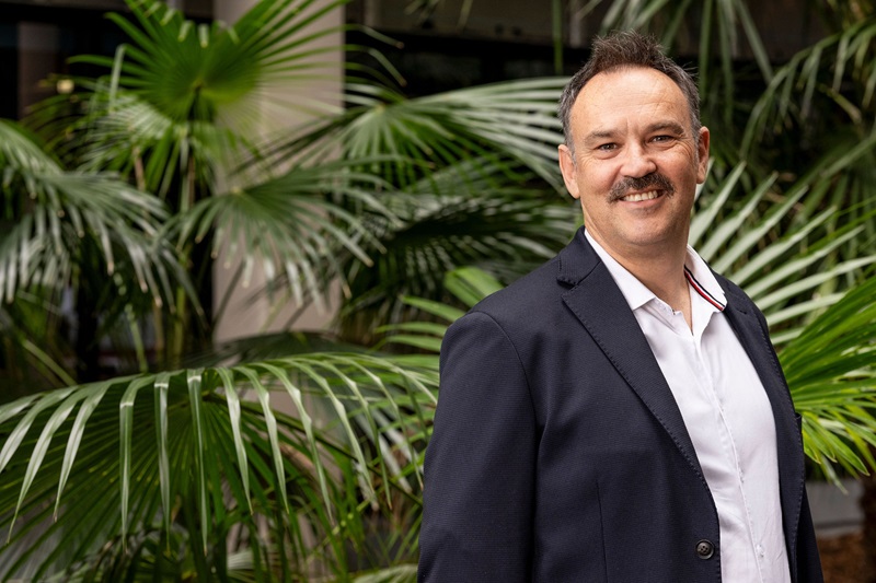 A man standing in front of ferns and wearing a suit, smiling at camera