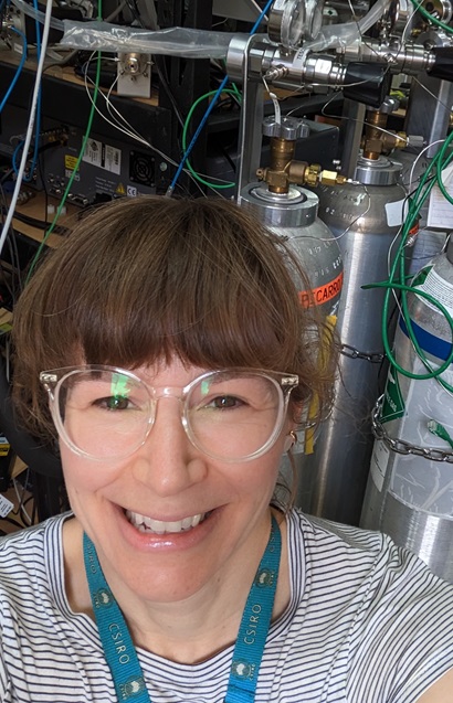 A female researcher wears glasses and smiles at the camera. She is standing in front of large air tanks containing air samples. 