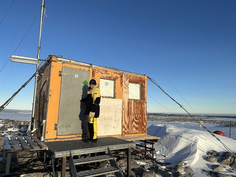 A researcher in a yellow jacket and pants stands outside a small hut, with the icy backdrop of Antarctica seen behind.