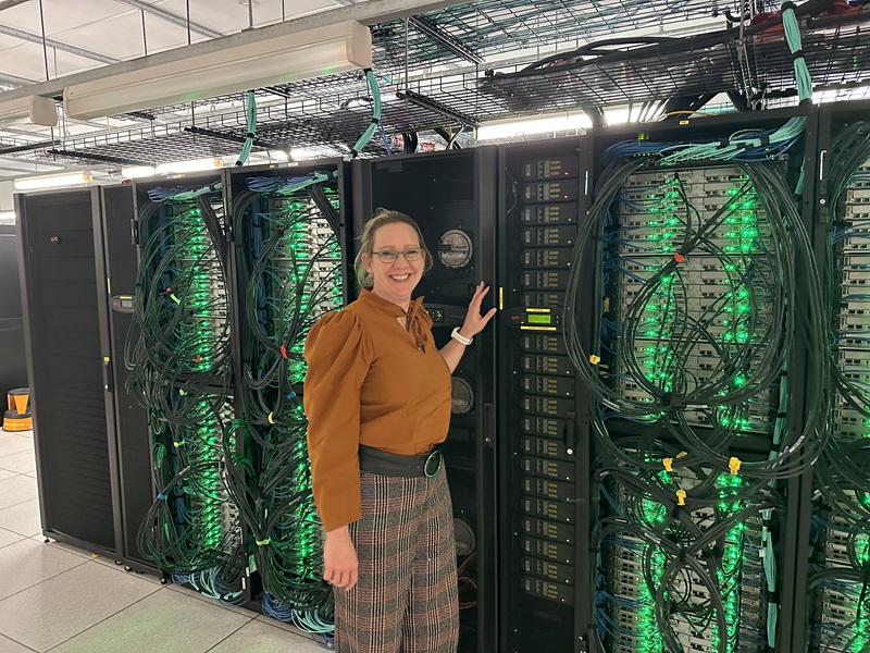 A woman wearing a mustard colour top and glasses, standing in front of a series of super computers