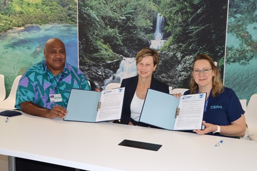 Three people seated at a table. A man and a woman on either end are smiling and holding up a signed contract. The person in the middle is also smiling warmly, and is wearing a suit jacket.