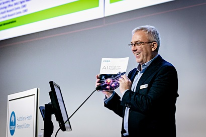 Man in blue suit holds up a report about AI whilst giving a speech behind a lectern. 