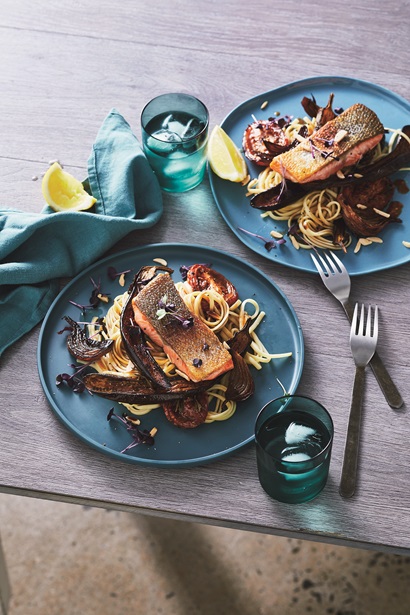 Two blue plates which contain soba noodles and a piece of crispy skin Salmon fillet on each. The plate are accompanied by two glasses of water with ice cubes in them and two forks