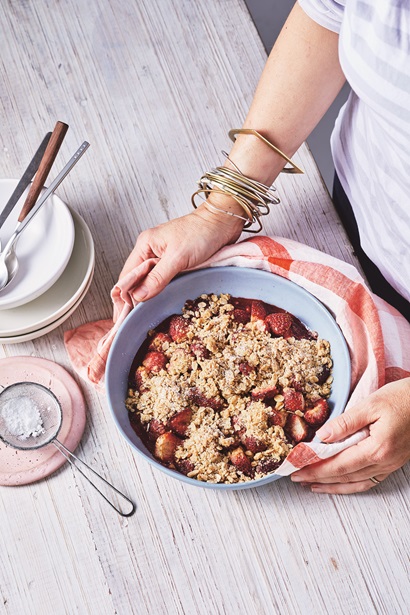 A white bowl filled with strawberries and an oat crumble that has been baked. A person is holding the bowl with a red and white checked tea towel. The person has a set of bangles on her wrist