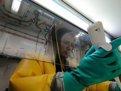 A scientist wearing a bright yellow fully contained biohazard suit, with their hands inside a fume hood, working with a pipette.