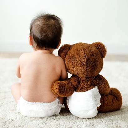 A baby sitting upright with its back to the camera in a nappy, next to the baby sits a teddy bear, upright also and with a nappy too, the teddy bear is leaning into the baby and has its arm around the baby in a caring way.