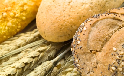 Three loaves of uncut bread are sitting on top of stalks of wheat. From left to right the loaves are a white loaf, a multigrain loaf and a wholemeal loaf coated in poppy and sesame seeds  