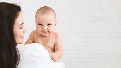 A laughing baby is looking over their mum's shoulder. Mum is wearing a white shirt and has long brown hair, she is smiling