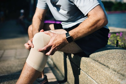 Person seated on bench while holding knee with visible strapping indicating knee pain.