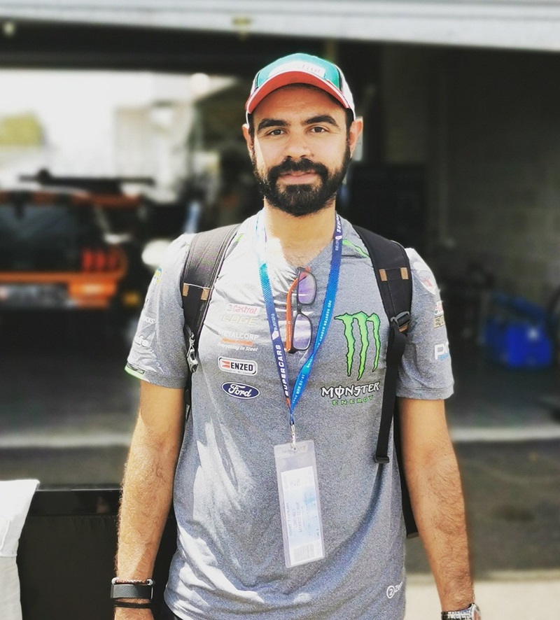 Photograph of a man wearing a T-shirt and baseball cap, with a lanyard around his neck. In the background is some racing cars. 