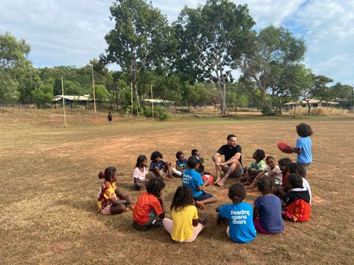 An adult man sits in a circle joined by Indigeous school children. One of the children is standing up talking to the man. They are sitting on grass which looks quite dry. There are trees which look like gums and a school building in the background.
