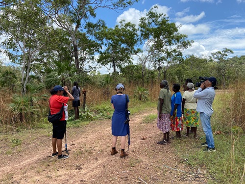 A group of Indigenous community members are talking to camrea. A cameraman wearing jeans and denim is standing behind the people. They are on a path surrounded by gums and green grass. 