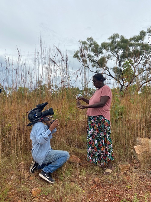 An Indigenous woman is standing in a field of long grass. She is holding what looks like a piece of bark and is explaining it to a camera man, who is recording her. She is wearing a pink top and long flowing floral skirt. He is wearing a denim top and denim jeans with sneakers.