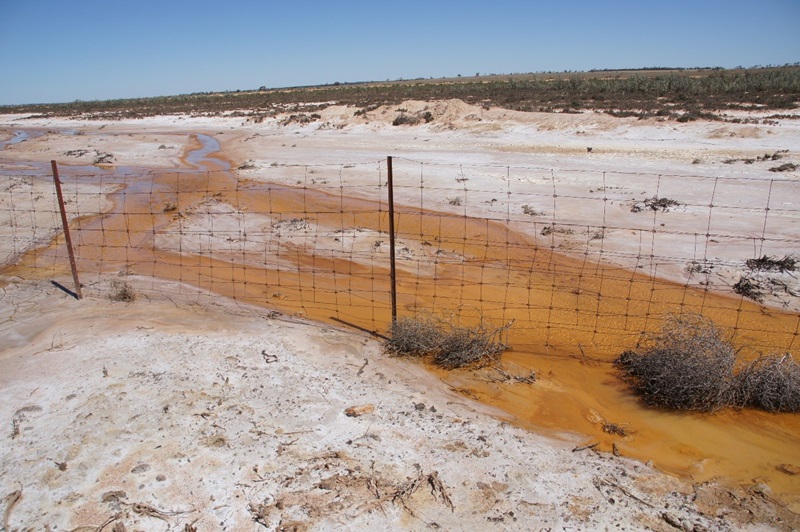 An acidic stream flowing across a barren environment, under a wire fence
