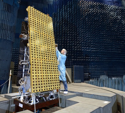 A man working on a golden metal rectangular satellite larger than the man inside a specialised chamber 