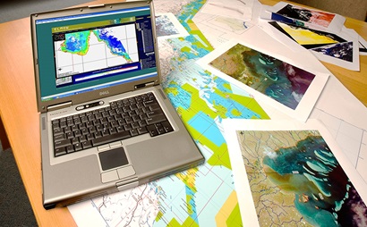 A desk with satellite maps spread across it and a laptop computer showing a satellite map of northern Australia on the screen
