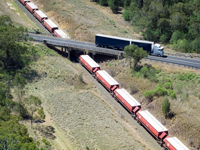 Picture of semi trailer travelling along a bridge with an image of freight train shown travelling on rail directly underneath the bridge.